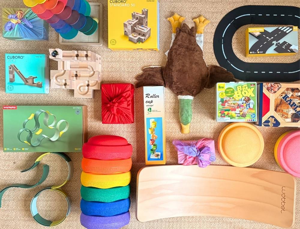 Premium toys arranged on a playroom rug, including a wooden CUBORO marble run, Stapelstein stepping stones,  Erzi wooden play food, WaytoPlay toy roads and small gifts wrapped in play silks