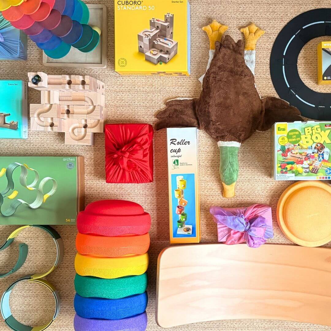 Premium toys arranged on a playroom rug, including a wooden CUBORO marble run, Stapelstein stepping stones,  Erzi wooden play food, WaytoPlay toy roads and small gifts wrapped in play silks