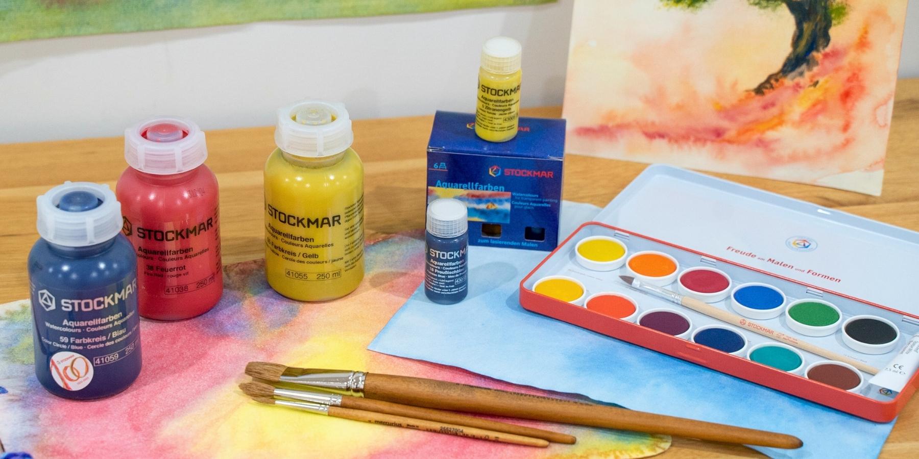 STOCKMAR Art supplies including paint bottles, a watercoloUr set, and brushes on a table with a painting in the background.