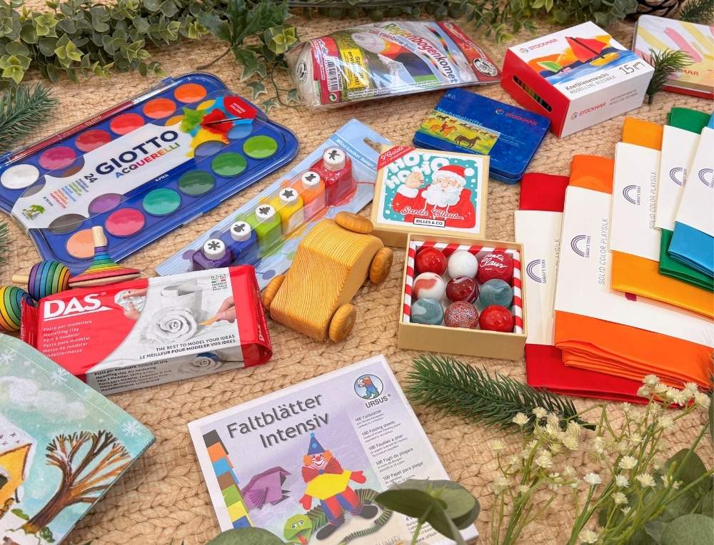 A selection of small toys and craft supplies spilling out of a Christmas stocking, including play silks, paints, wooden spinning tops, craft kits, glass marbles and other quality toys