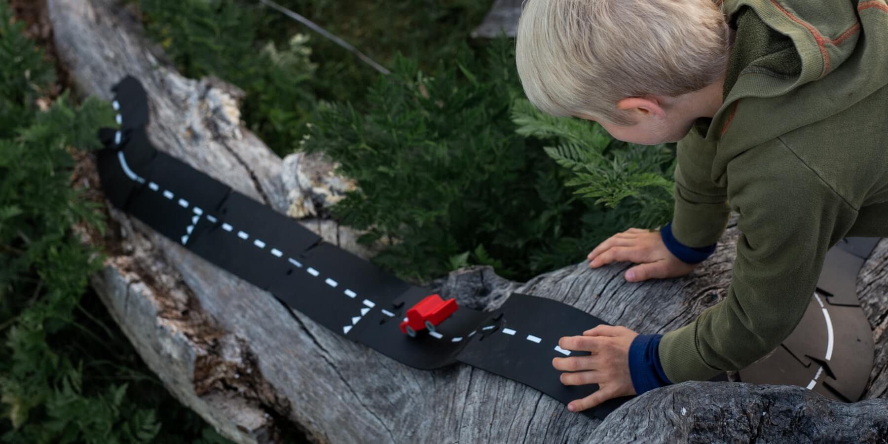 A child in an olive green jacket leans over a weathered log outdoors, playing with a black flexible WaytoPlay road track marked with white dashed lines and a small red vehicle.