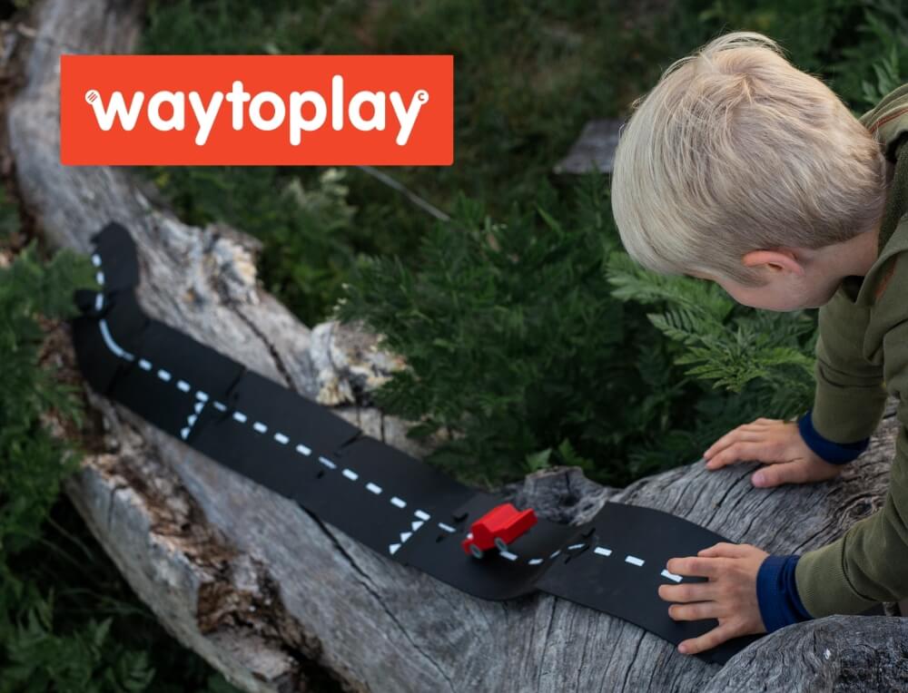 A child outdoors on a log using a WaytoPlay flexible road toy with a small red vehicle. The WaytoPlay logo is displayed in an orange box in the top left corner.