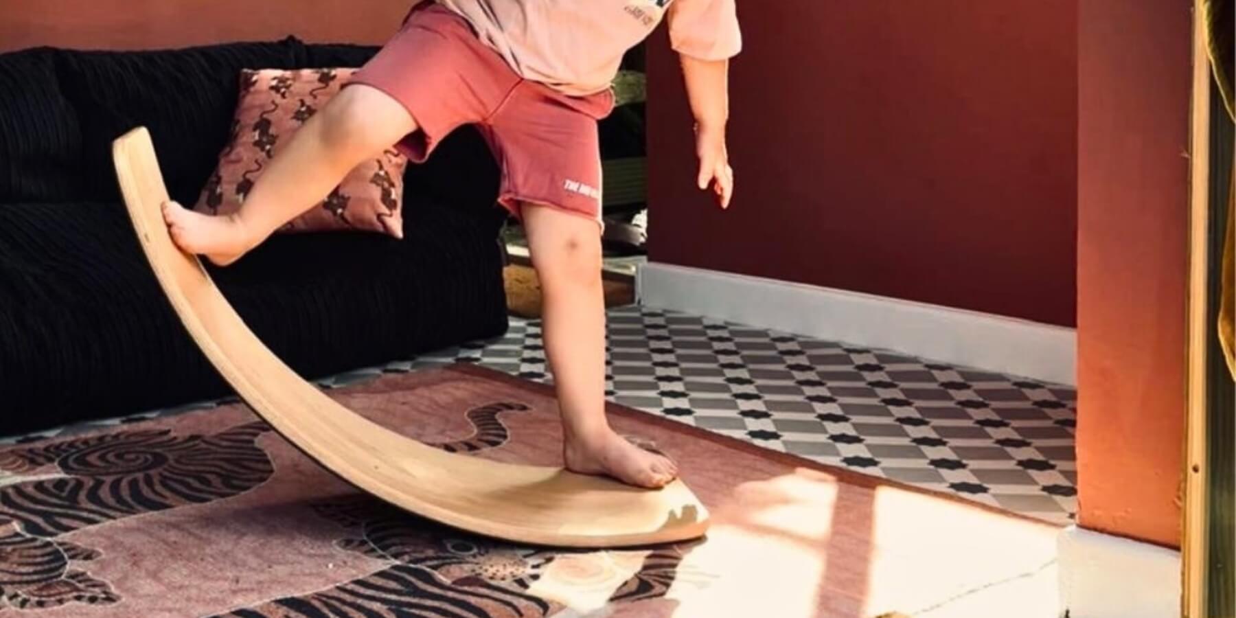 A child in pink shorts and a light top is balancing on a curved wooden wobbel board in a home interior with a patterned rug, dark sofa, and reddish-brown wall.
